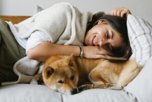 Cheerful brunette in comfy wear embracing fluffy adorable Shiba Inu dog while lying together on cozy couch in light living room